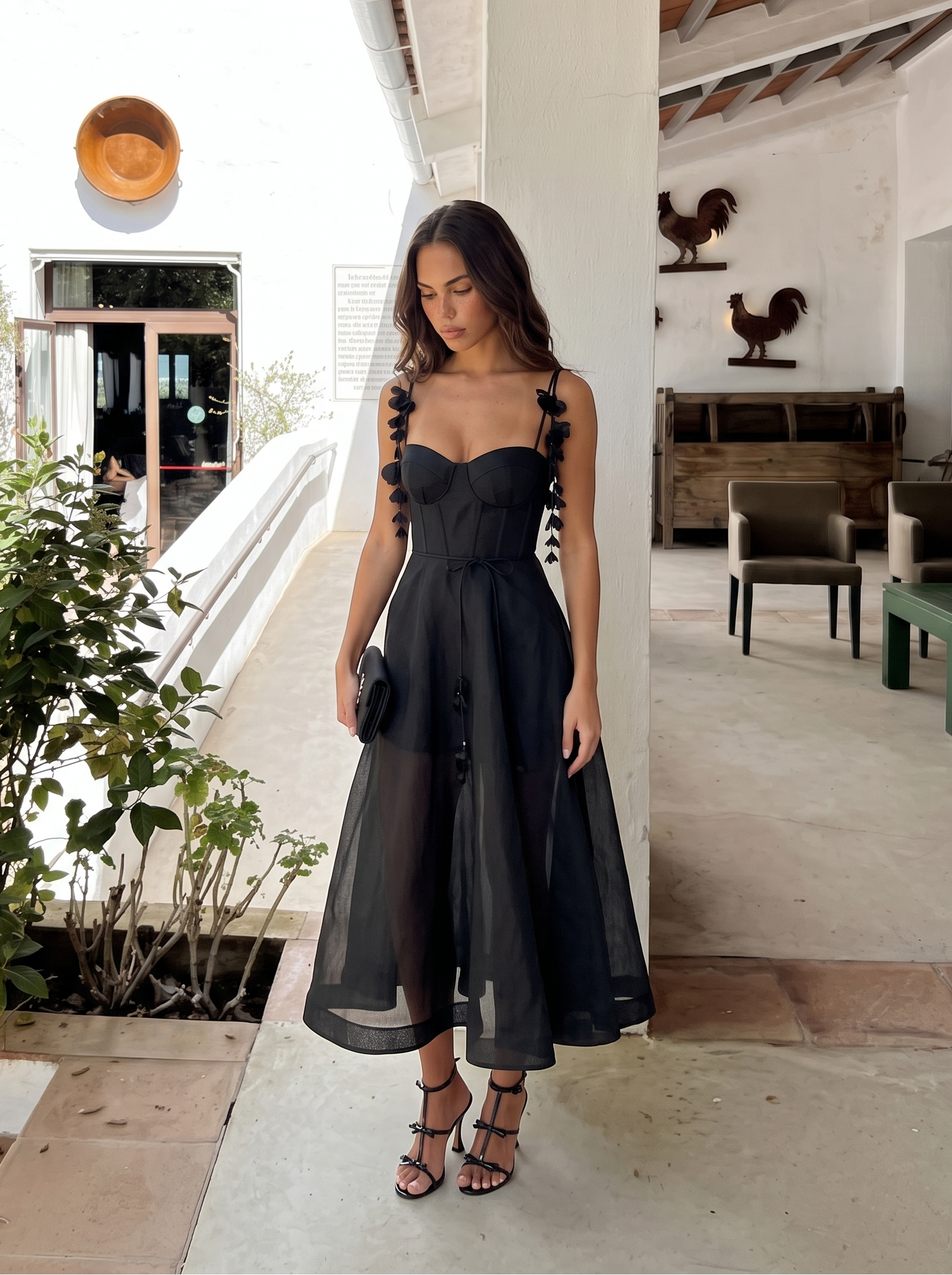 Woman in a black dress standing in a sunlit outdoor setting with plants and furniture.